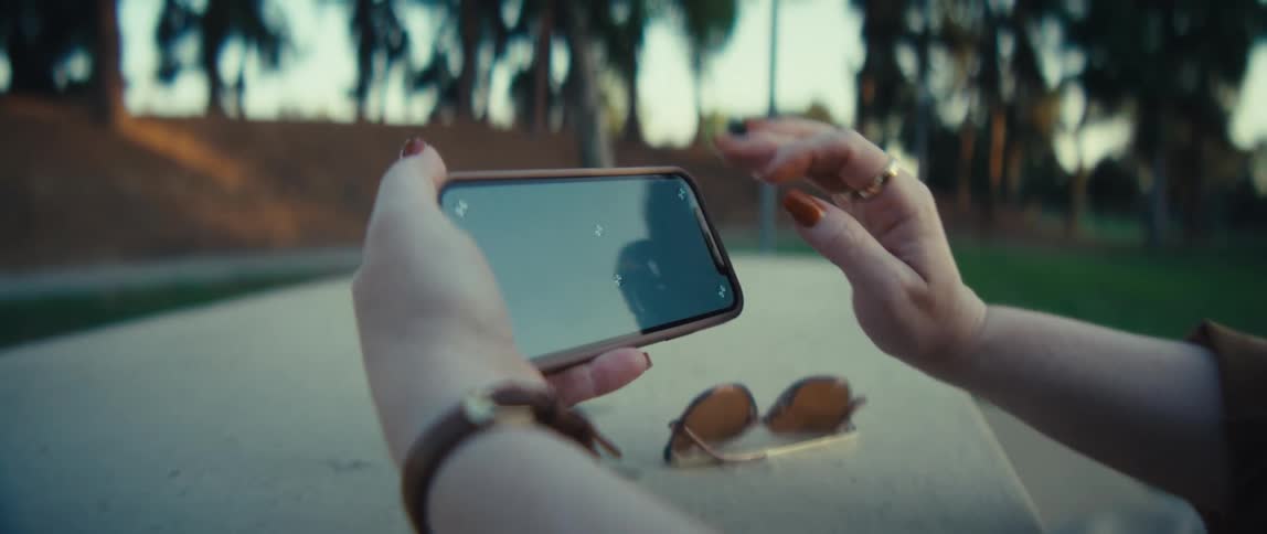Hands hold phone and sunglasses on outdoor surface with sky and greenery reflected
