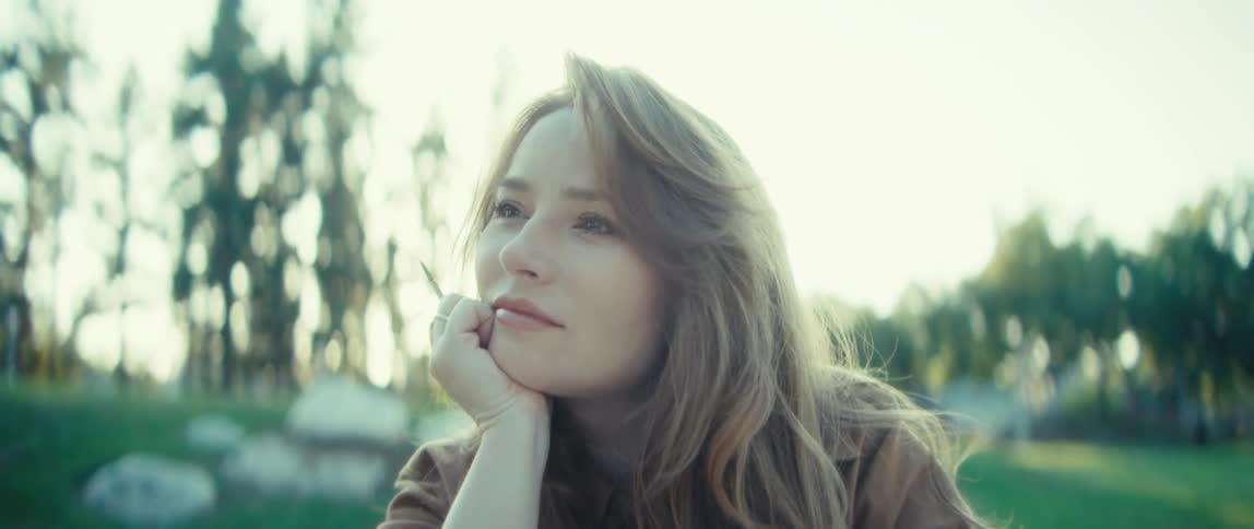 A young woman is sitting outside in a park wearing a brown jacket with sunlight filtering through the trees and highlighting her hair she is gently touching her face with her hand and looking up with a peaceful reflective expression