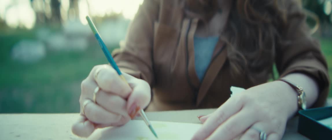 A woman paints a small watercolor design on paper at an outdoor table