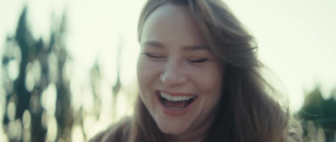 A young woman sits on a bench in a public park at midday watching a game on her phone her face lights up with excitement as she reacts to the action unfolding on the screen