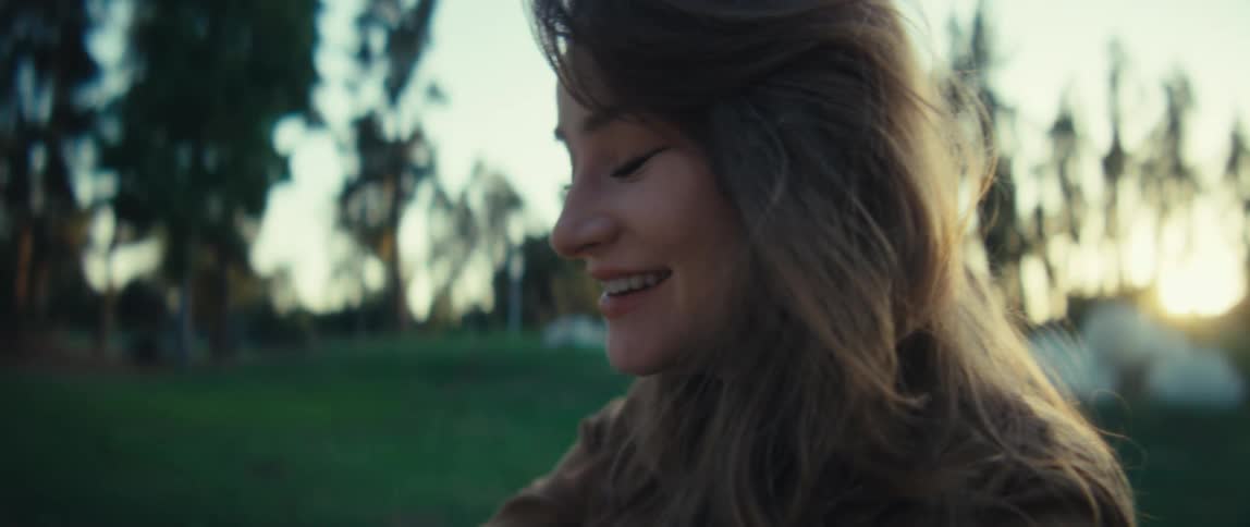 A closeup shot focuses on a persons hands as they tilt a smartphone that has a horizontal screen orientation the person appears to be smiling and laughing in the background which features blurred green trees and sunlight