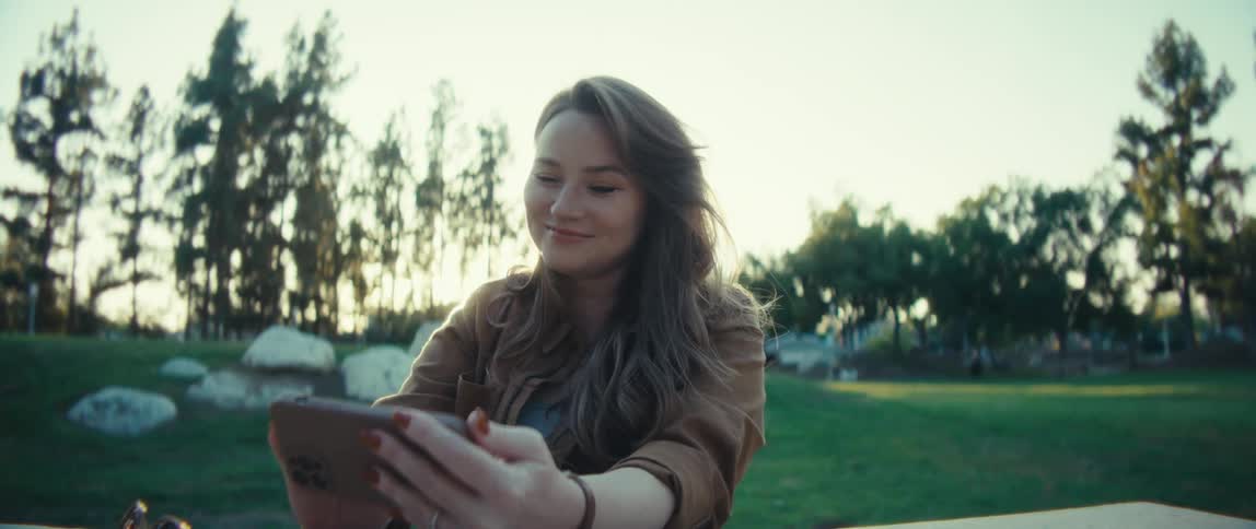 Close up of a young woman using a mobile phone while sitting at a table in an outdoor park at sunset