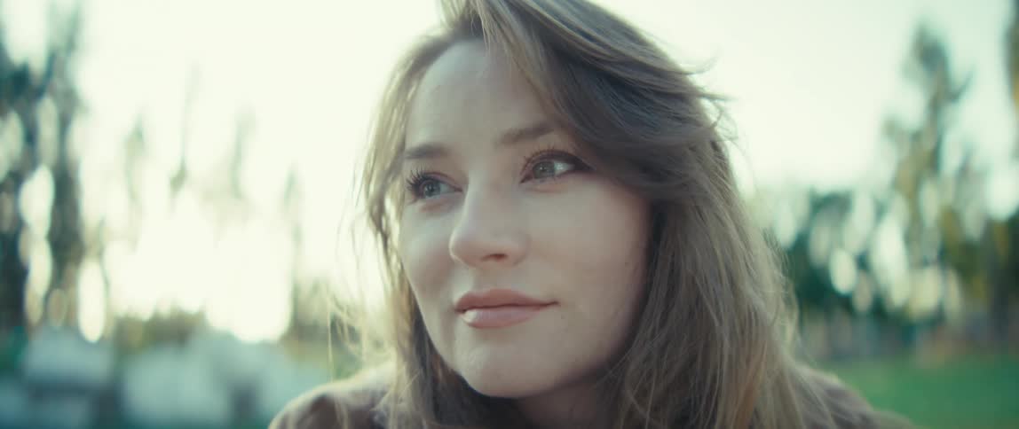 A young female artist sits on a bench in a public park at midday painting thoughtfully on a piece of paper as sunlight filters through the trees