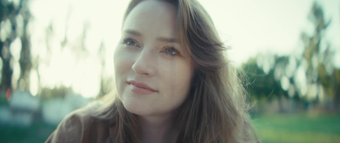 A young woman sits outside in a park with sunlight illuminating her face and hair she looks up with a gentle reflective expression her eyes conveying a peaceful feeling