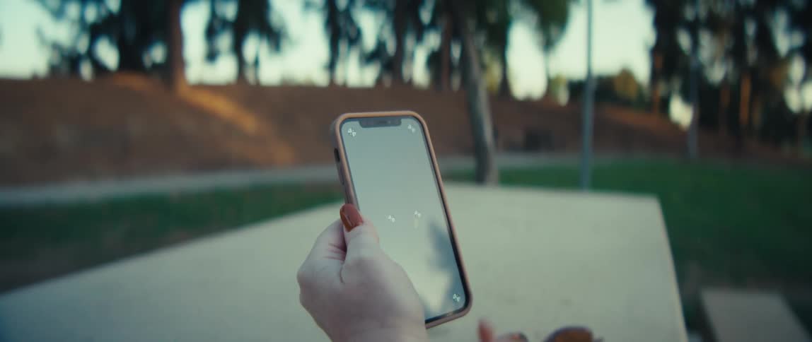 A closeup shot focuses on a persons hands as they scroll and interact with a smartphone outdoors the background is blurred showing sunlight streaming through trees in a park or similar green space