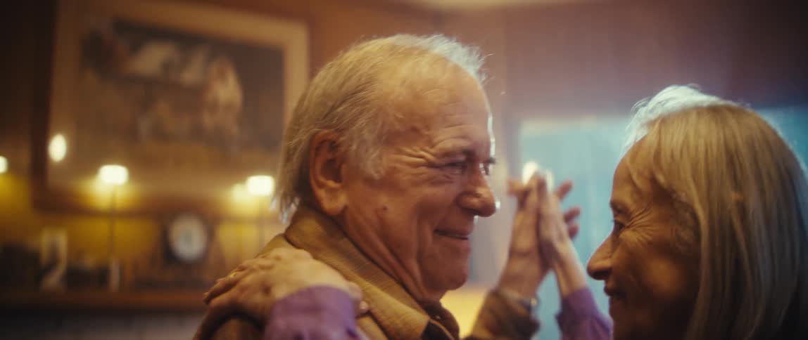 A closeup shot focuses on an elderly man with white hair and a tan shirt who is smiling and laughing the warm soft lighting in the indoor setting highlights his happy and engaged expression