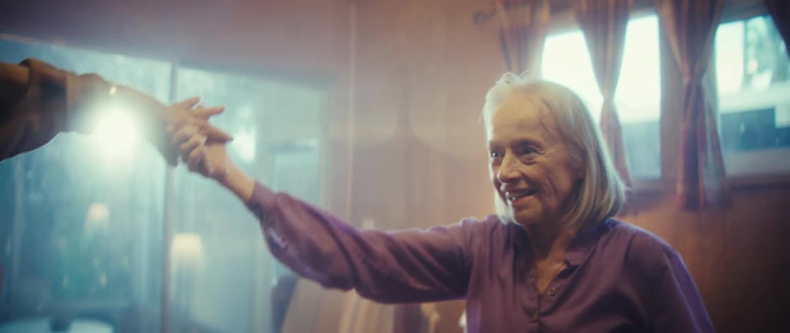 An elderly couple dances joyfully in their living room at midday smiling excitedly as they move together in a warm sunlit space