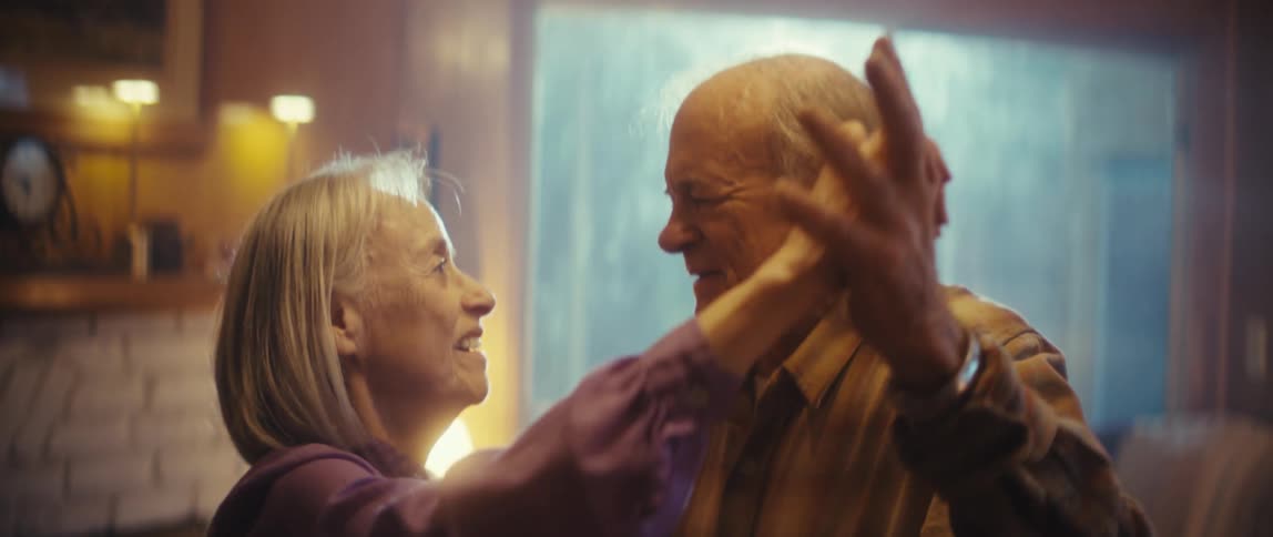 An elderly couple dances hand in hand in their living room smiling joyfully as they enjoy a fun lighthearted moment together