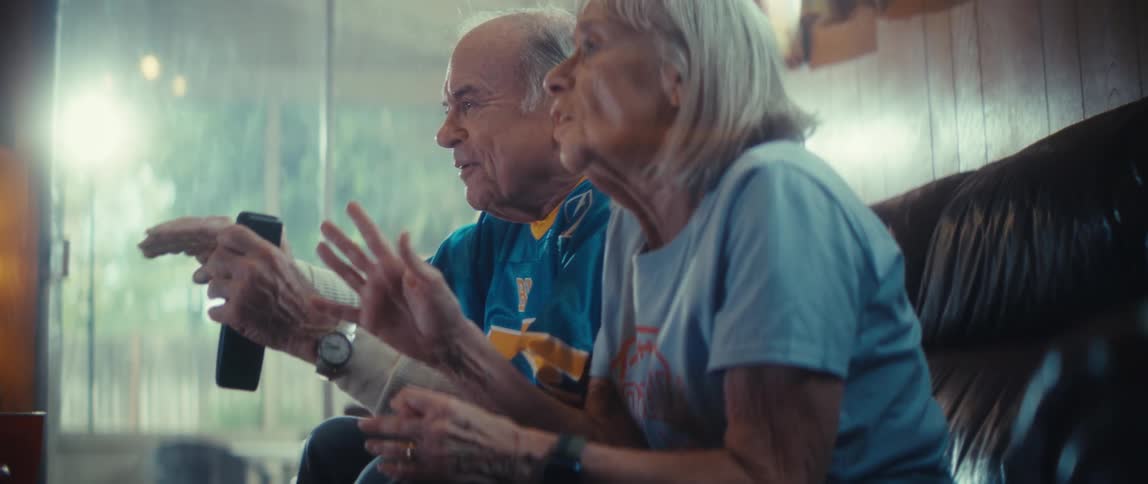 An elderly couple sits on the couch in their living room at midday watching a game with excited expressions as they cheer and react to the action unfolding on screen