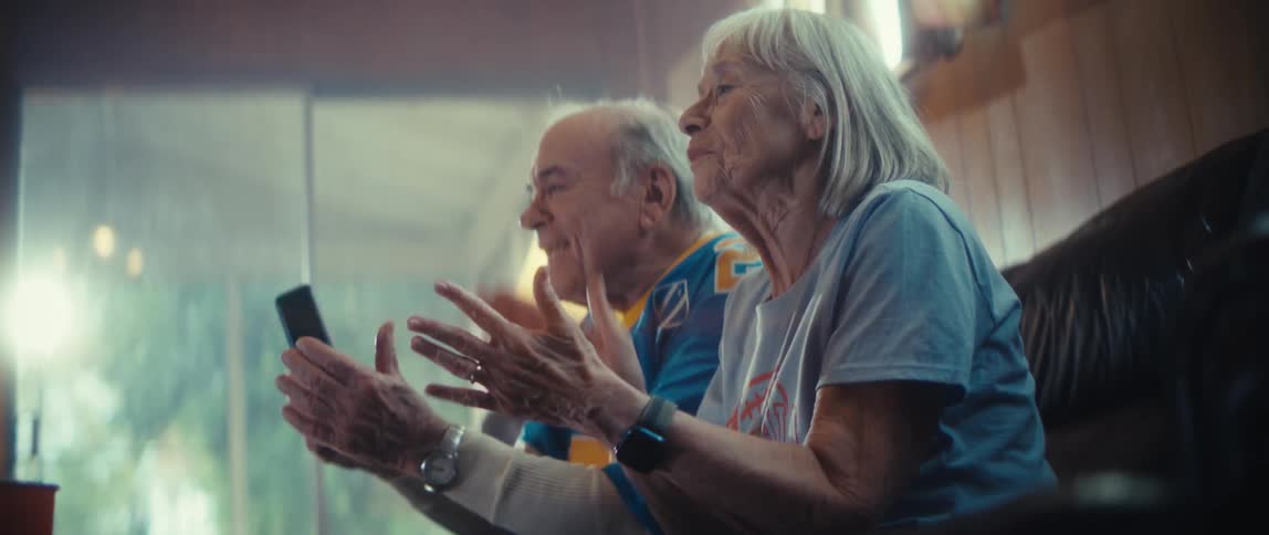 An elderly couple is clapping and moving as they sit on a couch