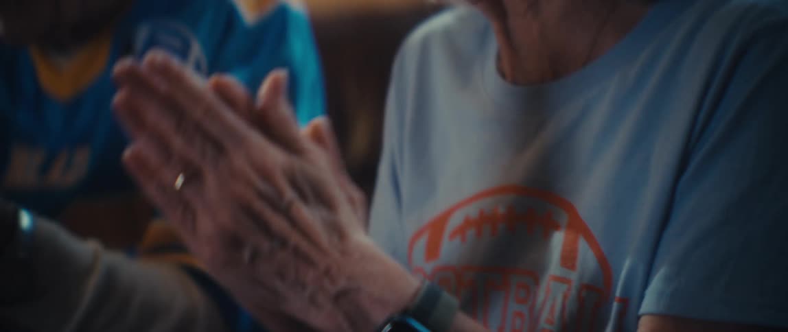 The video is a closeup of an elderly womans hands clasped together and gently rocking as she watches something offcamerashe is wearing a simple ring on her left hand and has a darkfaced smartwatch on her wrist and her expression suggests