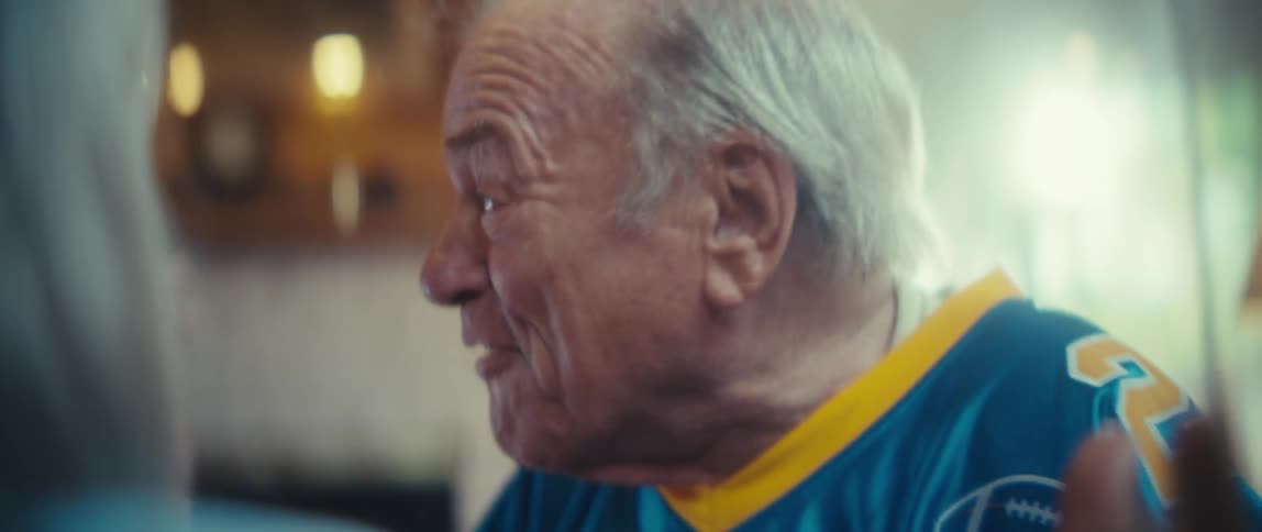 Elderly man with white hair laughs broadly while wearing blue sports jersey with yellow number