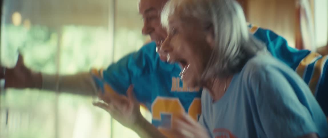 An elderly couple sits close together holding hands leaning forward with concerned faces then suddenly yelling with mouths wide open before enthusiastically hugging in an indoor setting