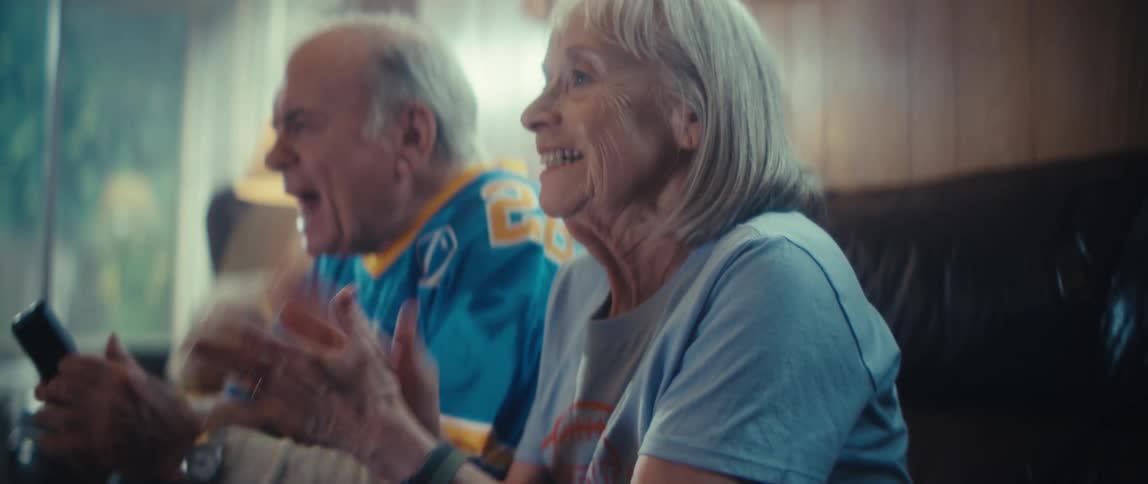 An elderly couple sits on a couch in their living room at midday cheering with excitement as they watch a game unfold their faces light up with joy sharing the thrill of the moment together