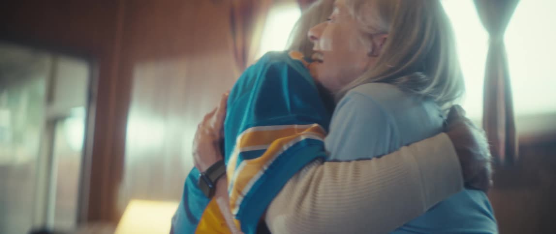 An elderly couple watches a game in their living room at midday bursting with excitement as they hug each other in celebration