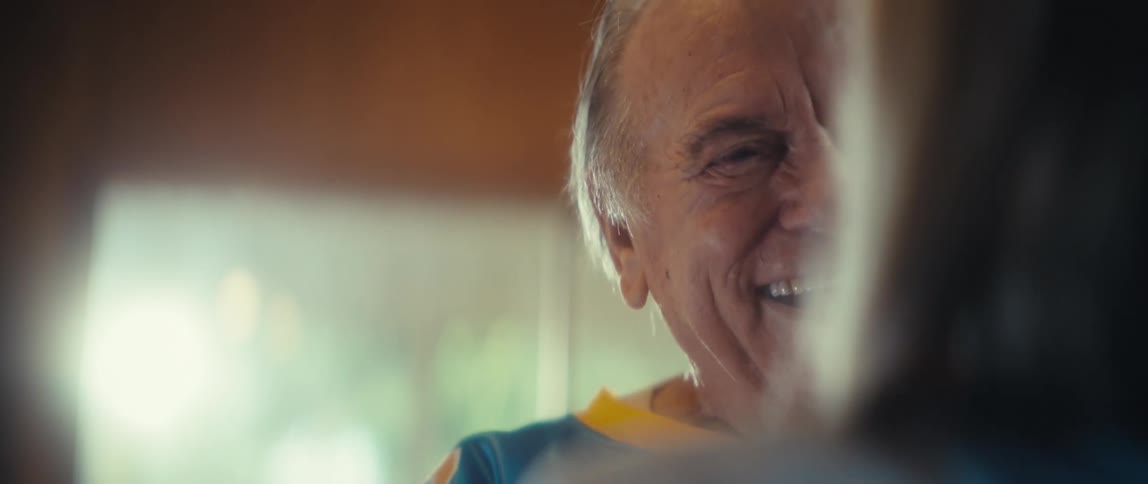 An elderly man in a blue football jersey laughs warmly and gives an elderly woman a joyful tight hug as they share a happy moment indoors