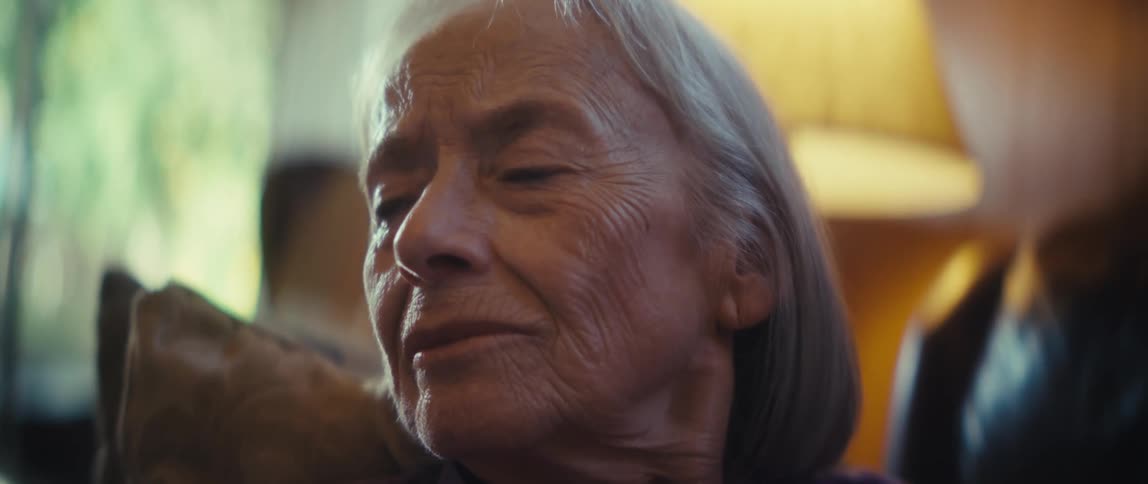 Elderly woman with gray hair wears purple top and looks to the side in indoor setting lit by a lamp