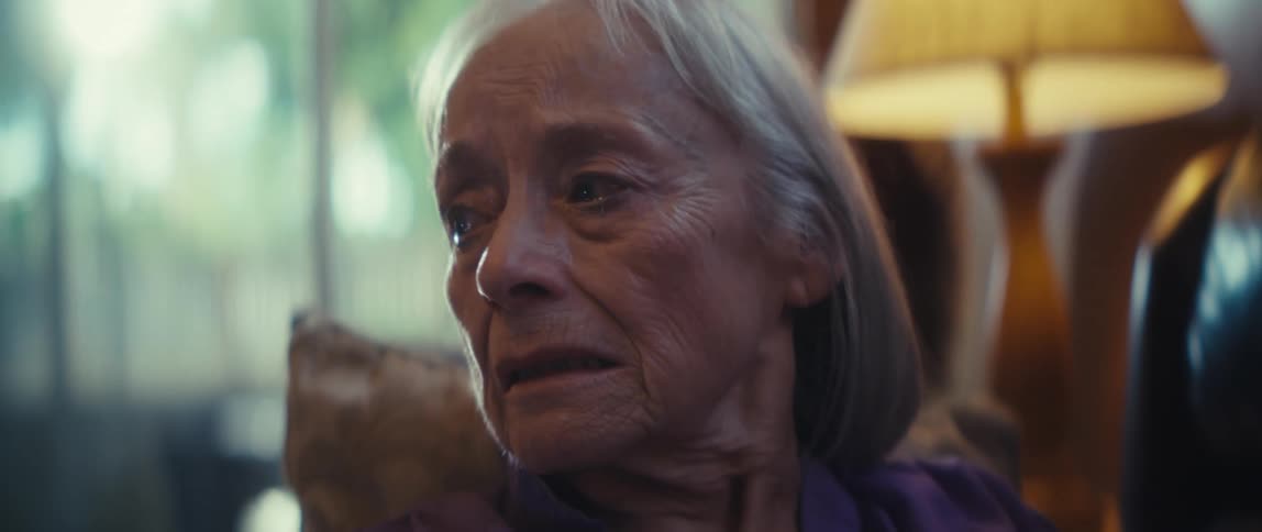 A closeup shot focuses on an elderly woman with white hair and a serious pensive expression looking upwards and offcamera the warm soft indoor lighting illuminates her face giving the scene a somber and contemplative mood