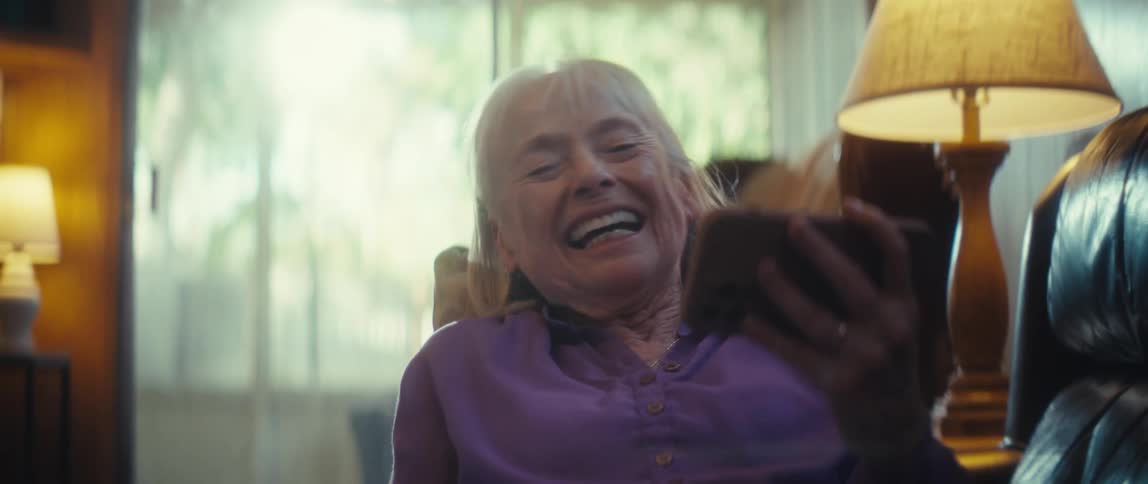 Elderly woman laughing with a phone in hand in a room lit by natural light and a lamp