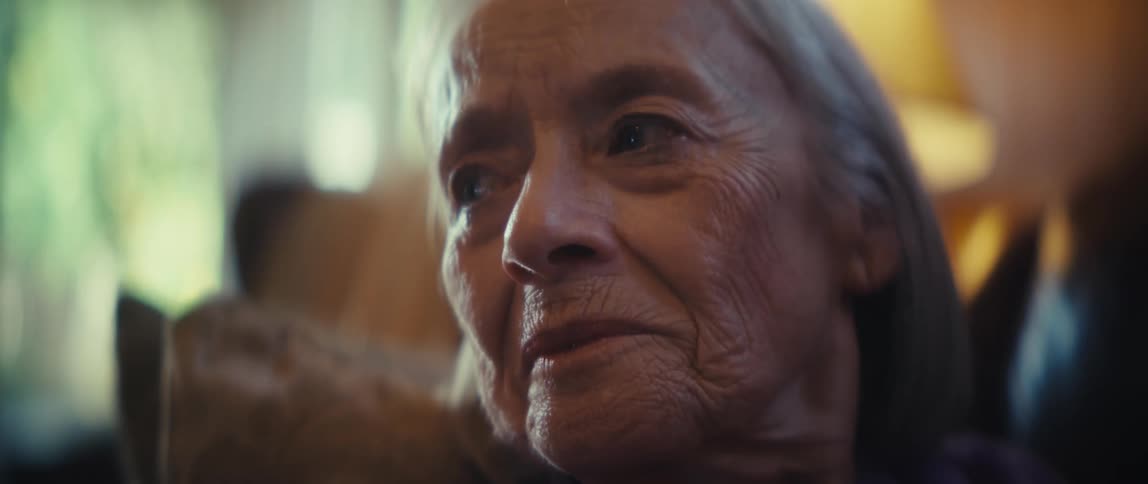 Elderly face with wrinkles and side gaze appears prominently under soft light and blurred background