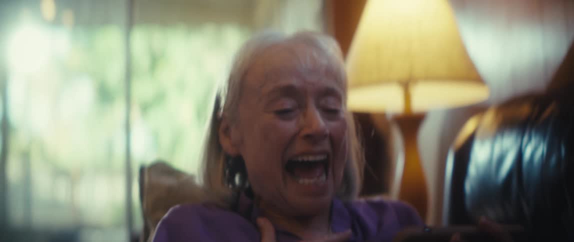 A closeup shot focuses on an elderly woman with white hair and a lavender shirt sitting in a chair and looking at her phone she is smiling broadly and laughing heartily suggesting she is enjoying something on the device