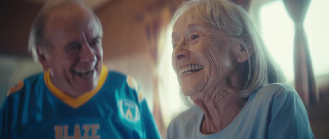 An elderly female claps with excitement as she talks to an elderly happy male in a well lit living room