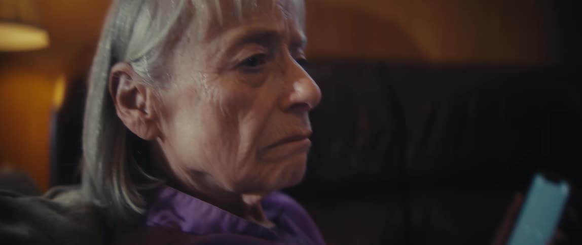 A silver haired older woman sits on a couch in warm lamplight holding a smartphone looking sideways with a nervous expression