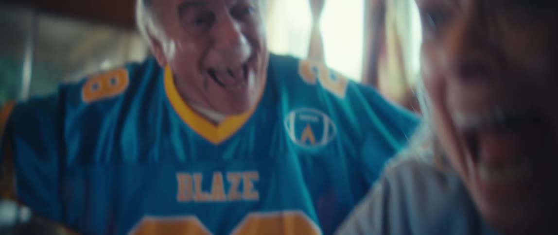 An elderly couple sitting on a couch reacts intensely to something on television by yelling raising their fists pointing the remote control and then embracing and laughing in celebration in a living room