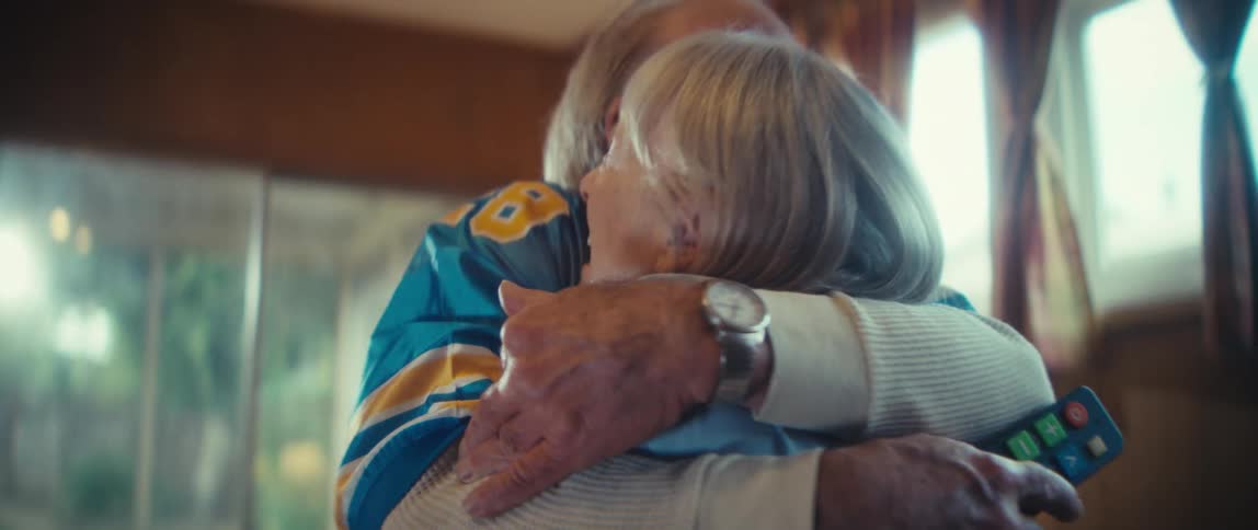 An elderly couple sitting on the couch cheering for their team then standing up celebrating and hugging each other