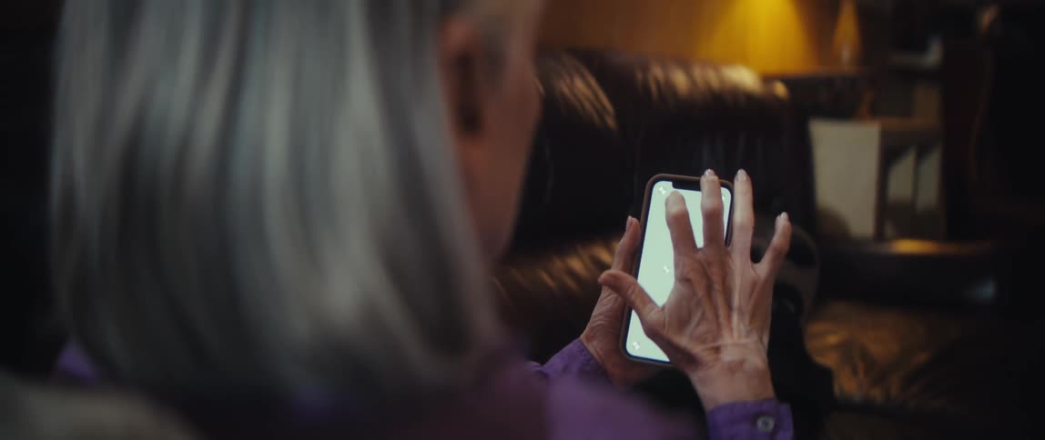 Elderly woman holds smartphone with light interface while seated on dark leather couch