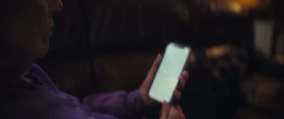 Elderly woman seated on couch looks at device with lit screen in lowlight setting