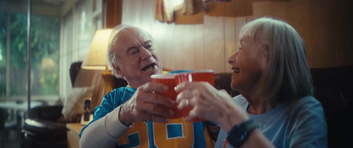 Elderly couple toasts with red cups in woodpaneled room with cozy lighting