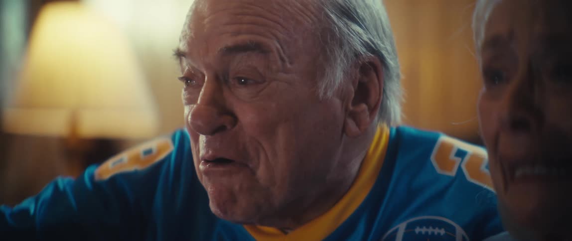 An elderly woman clasps her hands nervously before she and a man in a football jersey erupt in joyous celebration shouting and laughing with excitement and relief