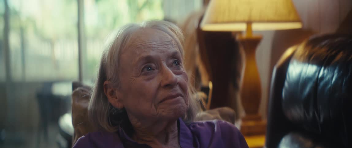 Elderly woman in purple shirt interacts with phone while seated on couch lit by natural light and lamp