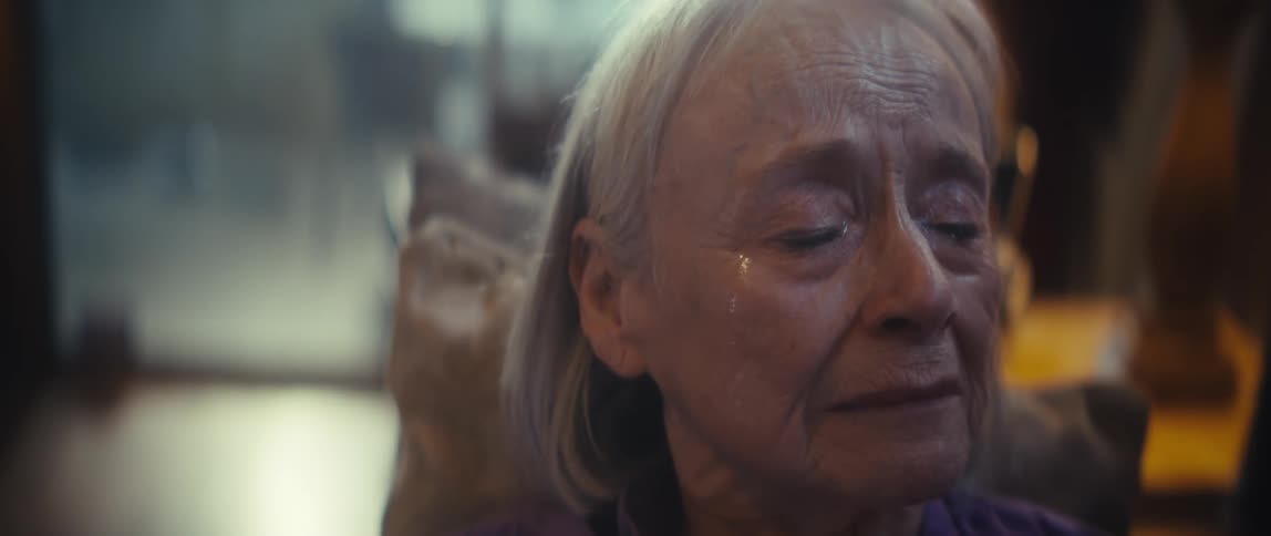 A closeup shot focuses on an elderly woman with white hair and a lavender shirt as she looks up and slightly offcamera with a contemplative expression her face is illuminated by warm soft indoor lighting highlighting a look of deep thought or perhaps