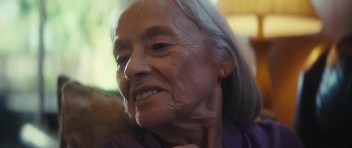 Elderly woman holds phone with brown case wearing purple shirt and ring on hand in softly lit setting