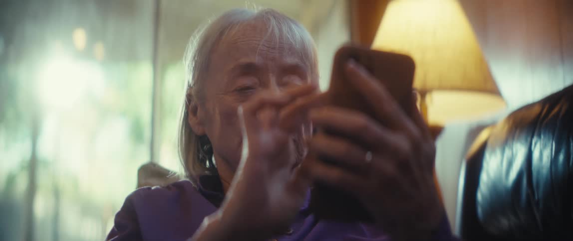 An elderly woman holds a mobile phone and swipes on the screen with one hand while sitting in a lamplit indoor space