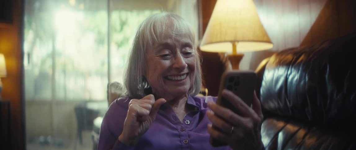 An elderly woman lies comfortably on the couch in her living room at midday smiling warmly as she scrolls through her mobile device she waves cheerfully during a video call engaged in a lively discussion