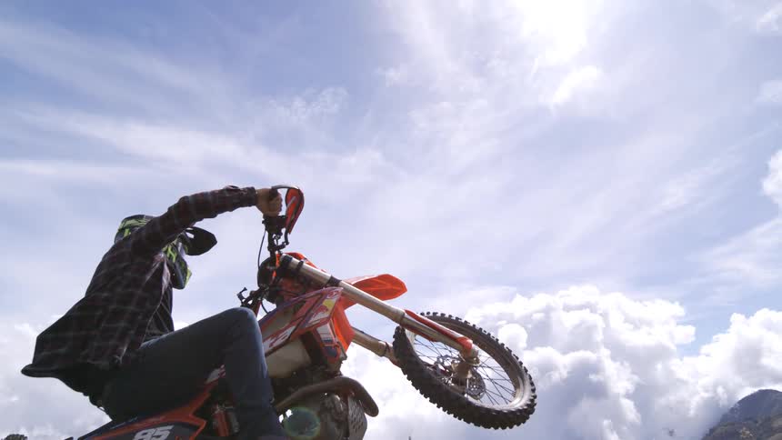 A motorcyclist lifts the front wheel on a mountain trail beneath a cloudy sky