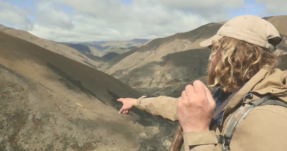 Man with backpack and walking stick points toward mountainous landscape under partly cloudy sky
