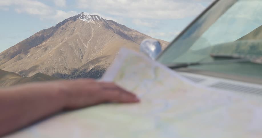 A persons hand rests on a paper map spread out across the hood of a vehicle with a large mountain dominating the background under a partly cloudy sky