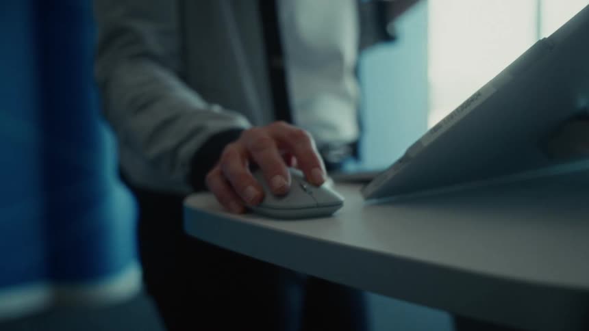He video shows a closeup of a persons hand using a grey computer mouse on a lightcolored deskthe person is wearing a lightgrey jacket and appears to be interacting with a computer screen or tablet that is angled upward on the desk