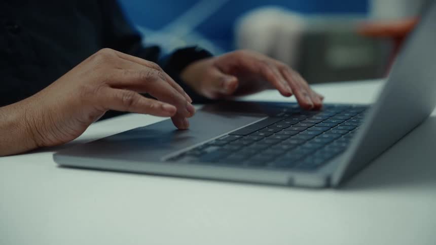 Fingers type on laptop keyboard over white surface with blurred blue wall in background