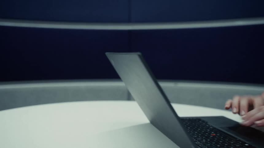A person wearing a green top and a watch opens a black laptop on a white table surface and immediately begins typing on the keyboard in a modern room
