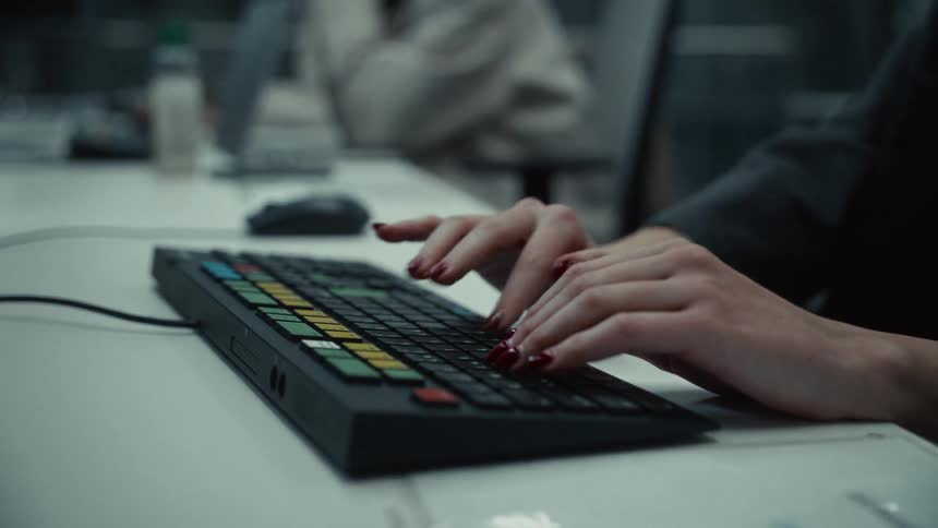 A medium closeup shot shows a persons hands with dark red nail polish rapidly typing on a black computer keyboard the keyboard is lowprofile with bright yellow and blue keys in a business setting