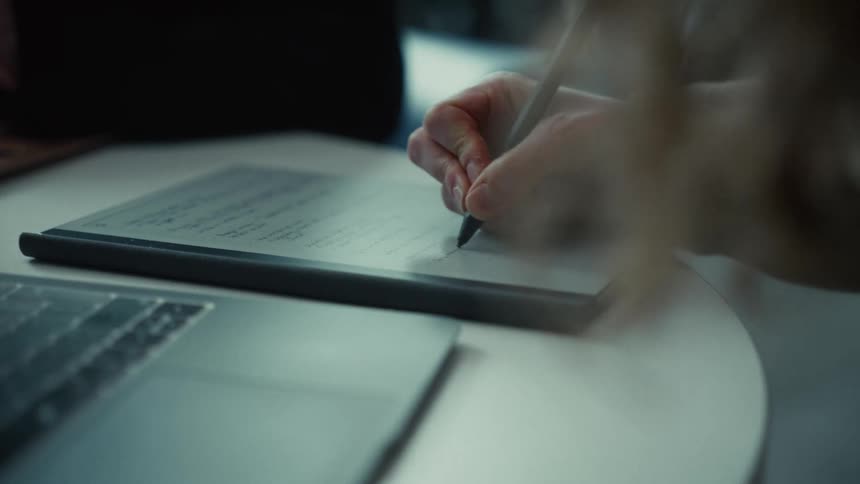 A woman sits behind a desk at midday writing with a pen on a document while a laptop rests nearby sunlight filters through the window casting a warm glow over her focused workspace