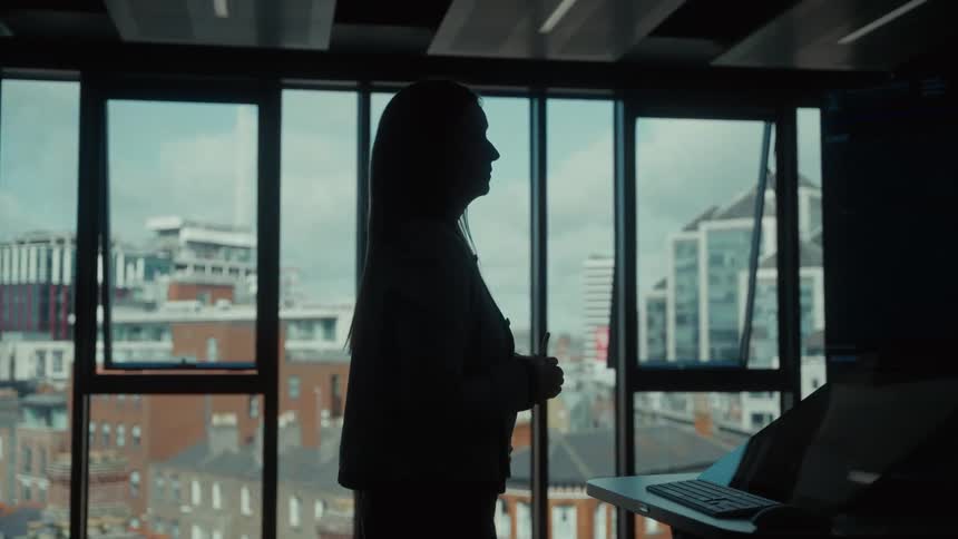The video shows the silhouette of a person with long hair standing in a modern office space facing a wall of large windows they appear to be giving a presentation or speaking as they gesture with their hands against the bright backdrop of a city skyline