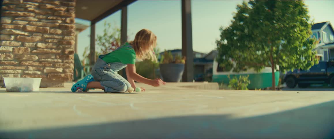 A young child wearing a green shirt and denim overalls is kneeling on a concrete patio or driveway in the sunlight they are actively engaged in drawing on the ground with a piece of colored chalk with a few other pieces of chalk scattered near them