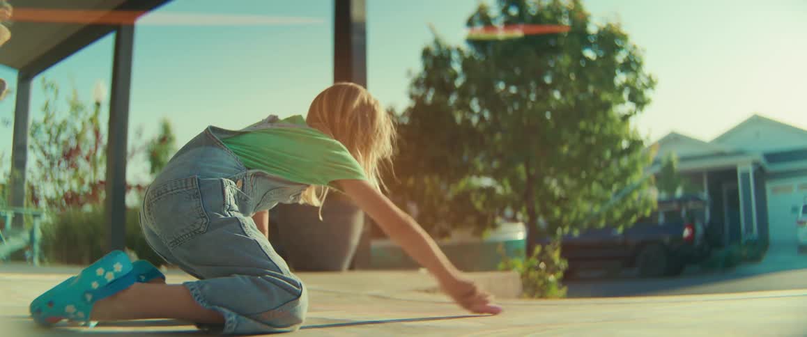 Child draws with chalk while kneeling on concrete in residential neighborhood with houses and trees in the background