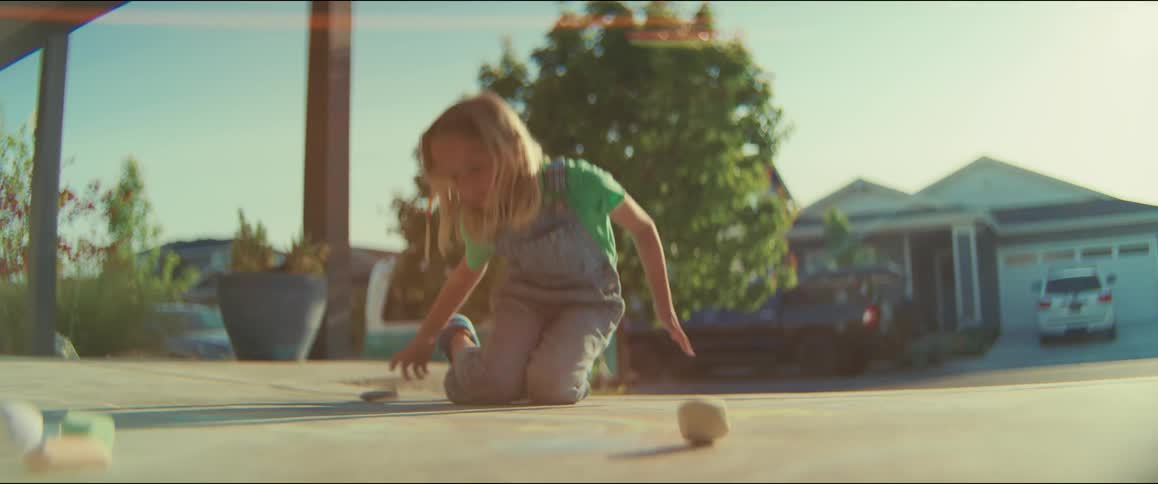 On a sunny suburban driveway a young girl in overalls draws with chalk before standing up and walking away